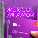 Cactos em fundo roxo, com as escritas em neon na parede "México Mi Amor". Em primeiro plano, uma mão segura o cartão roxo do Nubank