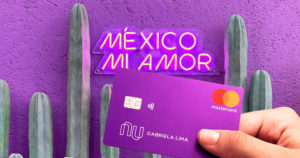 Cactos em fundo roxo, com as escritas em neon na parede "México Mi Amor". Em primeiro plano, uma mão segura o cartão roxo do Nubank